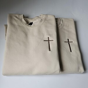 K&ouml;nnte beinhalten: Zwei beigefarbene Sweatshirts mit einem braunen gestickten Kreuz auf jedem. Die Sweatshirts sind gefaltet und &uuml;bereinander gestapelt.