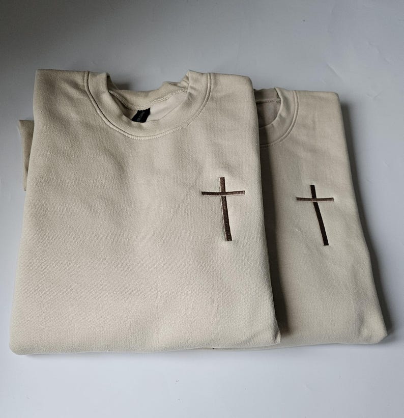 K&ouml;nnte beinhalten: Zwei beigefarbene Sweatshirts mit einem braunen gestickten Kreuz auf jedem. Die Sweatshirts sind gefaltet und &uuml;bereinander gestapelt.