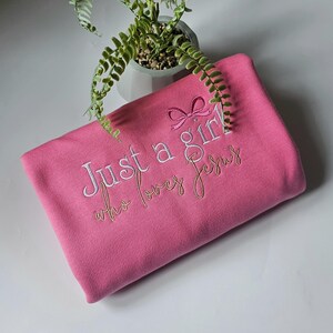 Op de afbeelding: Een roze sweatshirt met de geborduurde tekst "Just a girl who loves Jesus". De tekst is in witte en gouden draad, met een kleine strik boven het woord "girl". De sweatshirt is opgevouwen en ziet er zacht uit.