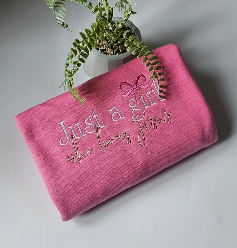 Op de afbeelding: Een roze sweatshirt met de geborduurde tekst "Just a girl who loves Jesus". De tekst is in witte en gouden draad, met een kleine strik boven het woord "girl". De sweatshirt is opgevouwen en ziet er zacht uit.