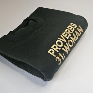 K&ouml;nnte beinhalten: Ein dunkelgr&uuml;ner Pullover mit goldener Stickerei, auf dem "Spr&uuml;che 31: Frau" steht.