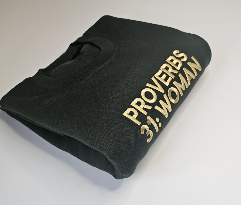 K&ouml;nnte beinhalten: Ein dunkelgr&uuml;ner Pullover mit goldener Stickerei, auf dem "Spr&uuml;che 31: Frau" steht.