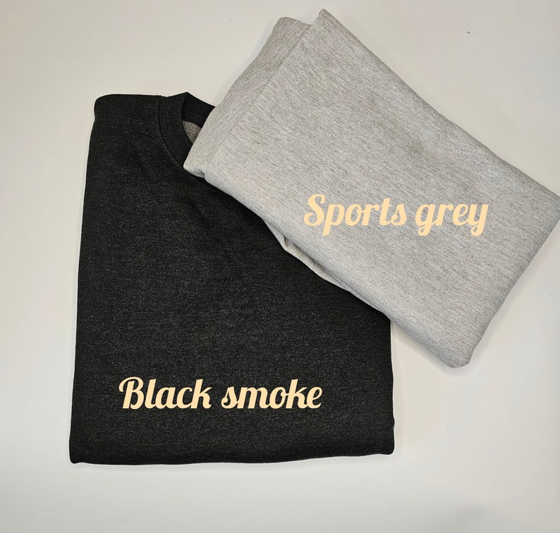 May include: Two folded sweatshirts, one black and one sports grey, are displayed on a white surface. The black sweatshirt has the text "Black smoke" in a cursive font. The sports grey sweatshirt has the text "Sports grey" in a cursive font.