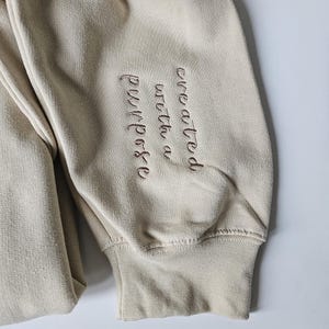 K&ouml;nnte beinhalten: Beigefarbener Sweatshirt mit einer braunen Stickerei, die "mit einem Zweck geschaffen" lautet.