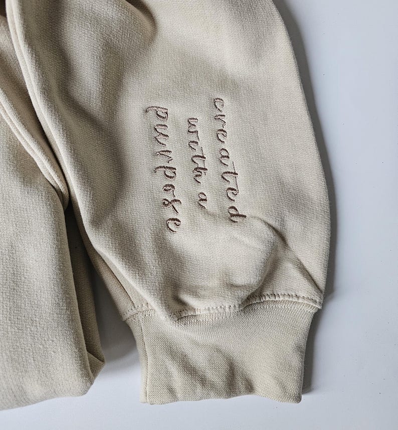 K&ouml;nnte beinhalten: Beigefarbener Sweatshirt mit einer braunen Stickerei, die "mit einem Zweck geschaffen" lautet.