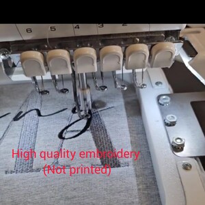 May include: Close-up of an embroidery machine in operation, stitching a design onto gray fabric. The machine's needles and white components are visible, with the text "High quality embroidery (Not printed)" in red.