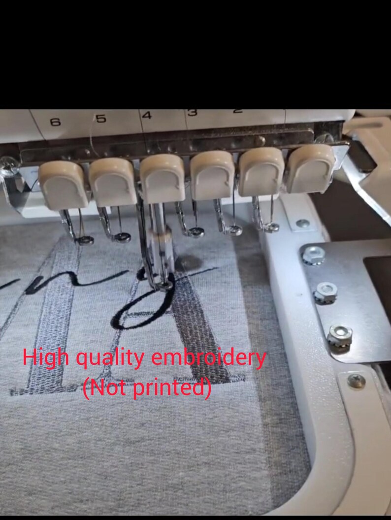 May include: Close-up of an embroidery machine in operation, stitching a design onto gray fabric. The machine's needles and white components are visible, with the text "High quality embroidery (Not printed)" in red.