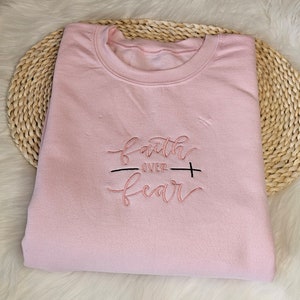 May include: A pink sweatshirt with the embroidered text "faith over fear" in a cursive font. The text is in a light pink color with a black line separating the words "faith" and "fear".