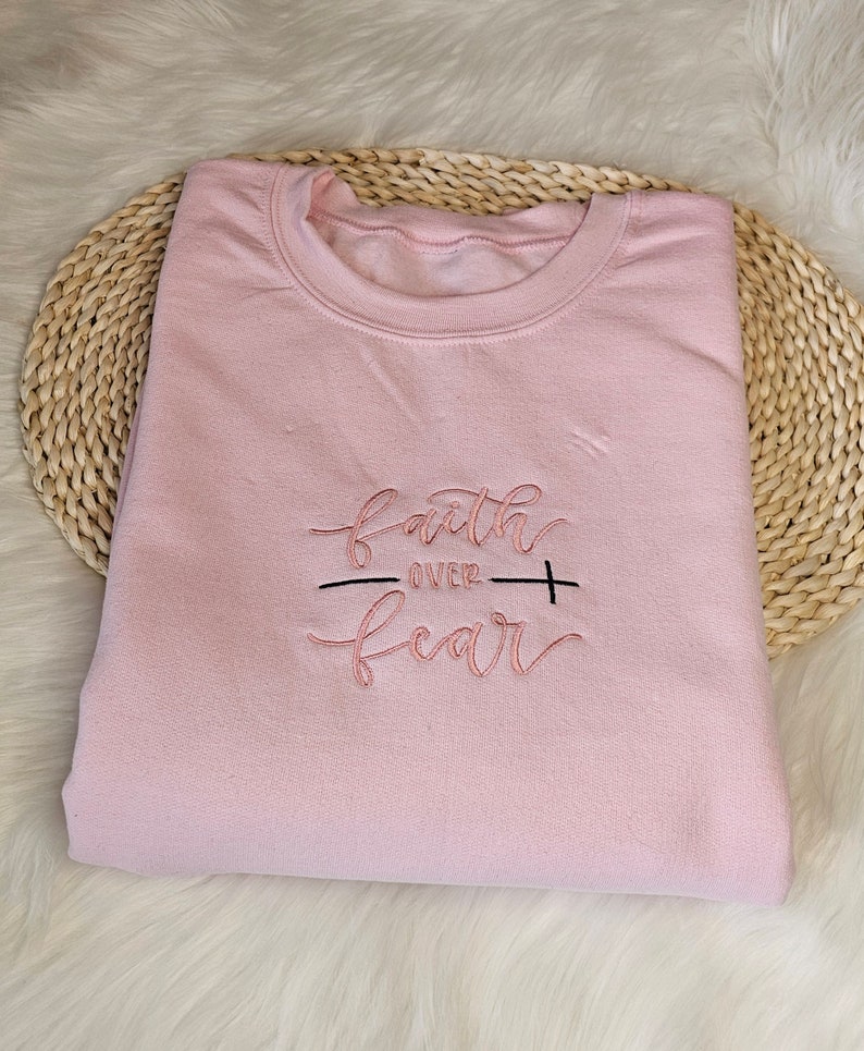 May include: A pink sweatshirt with the embroidered text "faith over fear" in a cursive font. The text is in a light pink color with a black line separating the words "faith" and "fear".