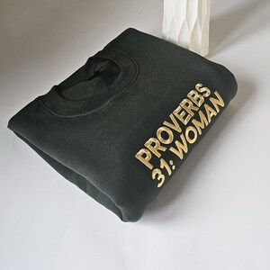 K&ouml;nnte beinhalten: Ein dunkelgr&uuml;ner Pullover mit dem Text "Spr&uuml;che 31: Frau" in goldener Stickerei.
