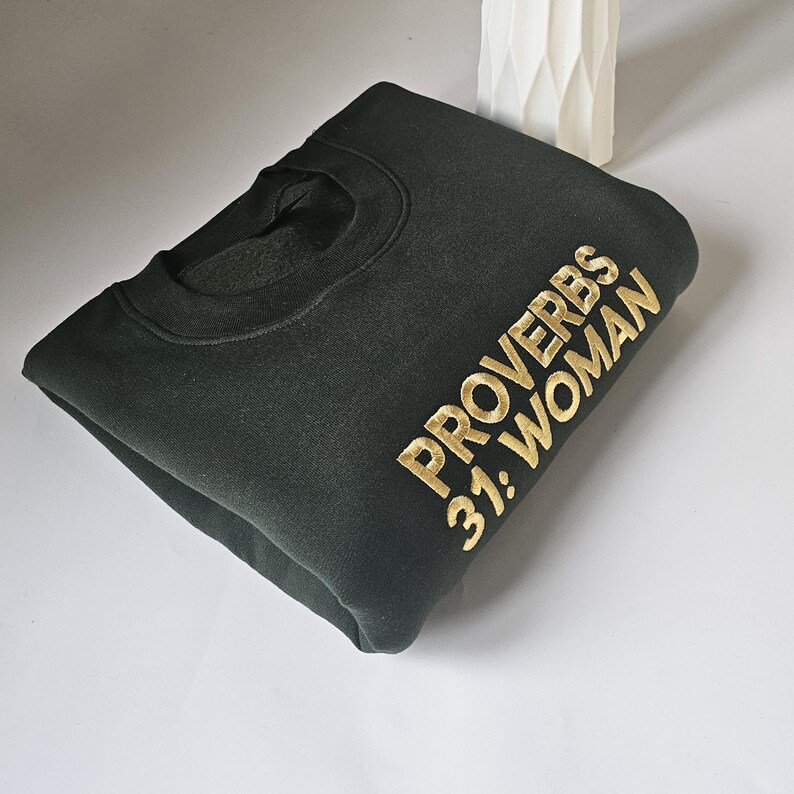 K&ouml;nnte beinhalten: Ein dunkelgr&uuml;ner Pullover mit dem Text "Spr&uuml;che 31: Frau" in goldener Stickerei.
