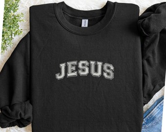Può includere: Felpa nera con la scritta "JESUS" in bianco, con un carattere in stile collegiale sul petto. La felpa è realizzata in un materiale morbido e ha un girocollo.