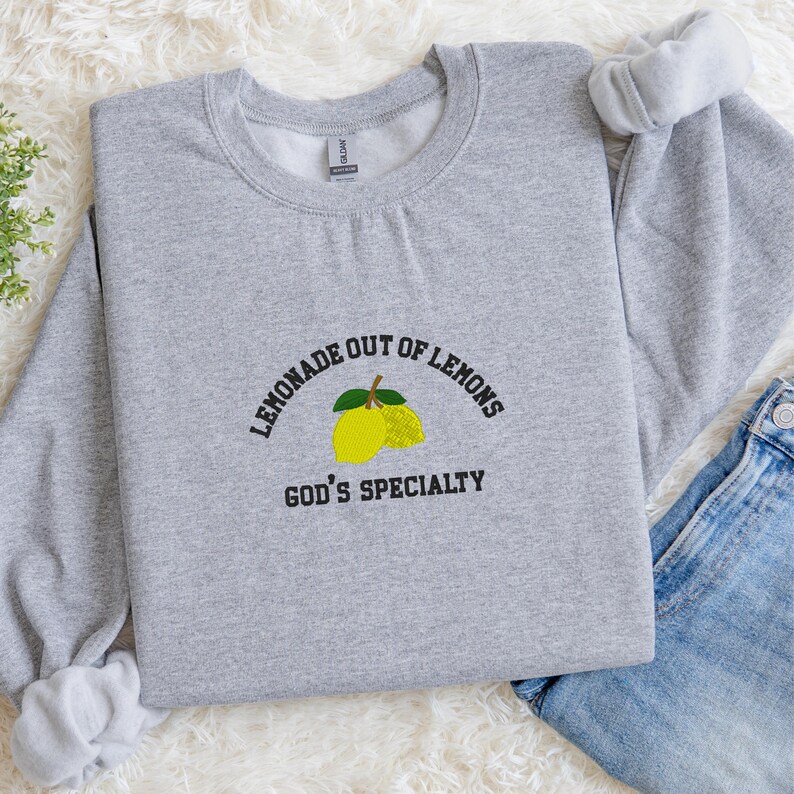 May include: A light gray sweatshirt with the embroidered text "LEMONADE OUT OF LEMONS" above a yellow lemon graphic and the text "GOD'S SPECIALTY" below. The sweatshirt has a crew neck and long sleeves.