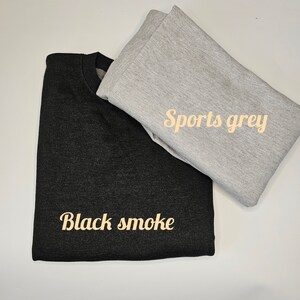 Op de afbeelding: Twee opgevouwen sweatshirts, &eacute;&eacute;n zwart en &eacute;&eacute;n sportgrijs, worden getoond op een wit oppervlak. Het zwarte sweatshirt heeft de tekst "Black smoke" in een cursief lettertype. Het sportgrijze sweatshirt heeft de tekst "Sports grey" in een cursief lettertype.