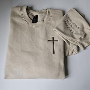 K&ouml;nnte beinhalten: Ein beiger Pullover mit einem braunen Kreuz, das auf der Vorderseite gestickt ist, und dem Text "geschaffen mit einem Zweck", der auf dem &Auml;rmel gestickt ist.
