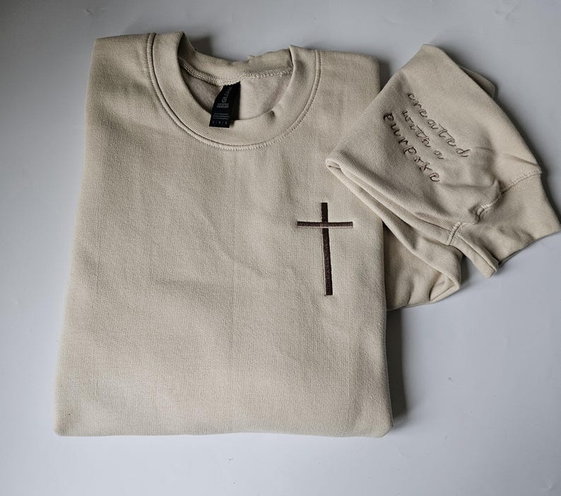 K&ouml;nnte beinhalten: Ein beiger Pullover mit einem braunen Kreuz, das auf der Vorderseite gestickt ist, und dem Text "geschaffen mit einem Zweck", der auf dem &Auml;rmel gestickt ist.