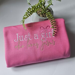 Op de afbeelding: Een roze sweatshirt met de geborduurde tekst "Just a girl who loves Jesus" in witte en gouden draad. Boven het woord "girl" is een roze strik geborduurd. Op de achtergrond staat een kleine plant in een pot.