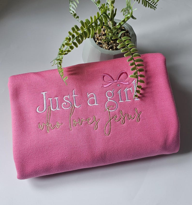 Op de afbeelding: Een roze sweatshirt met de geborduurde tekst "Just a girl who loves Jesus" in witte en gouden draad. Boven het woord "girl" is een roze strik geborduurd. Op de achtergrond staat een kleine plant in een pot.
