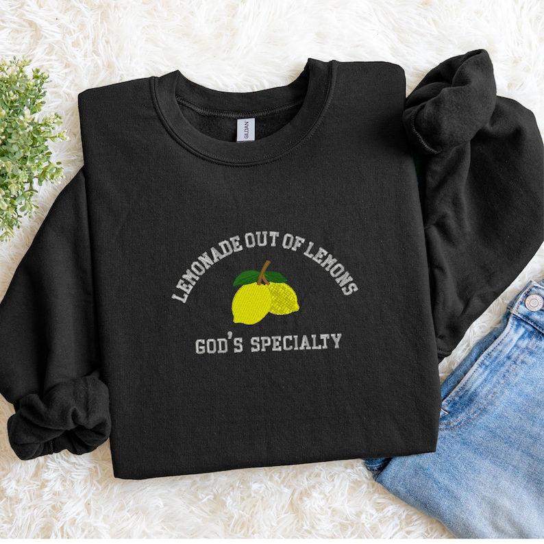 May include: Black crewneck sweatshirt with the embroidered text "LEMONADE OUT OF LEMONS" above a lemon graphic and the text "GOD'S SPECIALTY". The sweatshirt is folded on a white surface.