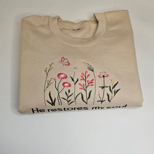 May include: A beige sweatshirt with embroidered flowers and the text "He restores my soul".