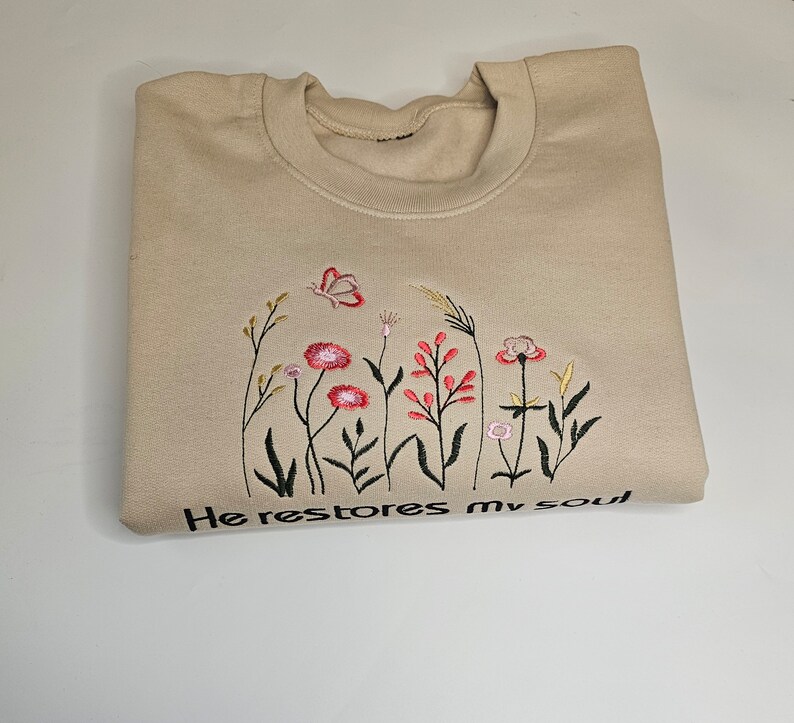May include: A beige sweatshirt with embroidered flowers and the text "He restores my soul".