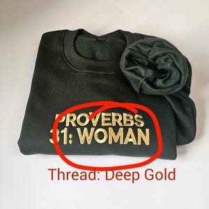 K&ouml;nnte beinhalten: Ein dunkelgr&uuml;ner Pullover mit dem Text "Spr&uuml;che 31: Frau" in goldener Stickerei. Der Text ist rot umkreist.