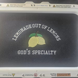 May include: Black fabric with embroidered text and a lemon design. The text reads "LEMONADE OUT OF LEMONS" above "GOD'S SPECIALTY". The design includes two yellow lemons and green leaves. The embroidery is framed by a white hoop.