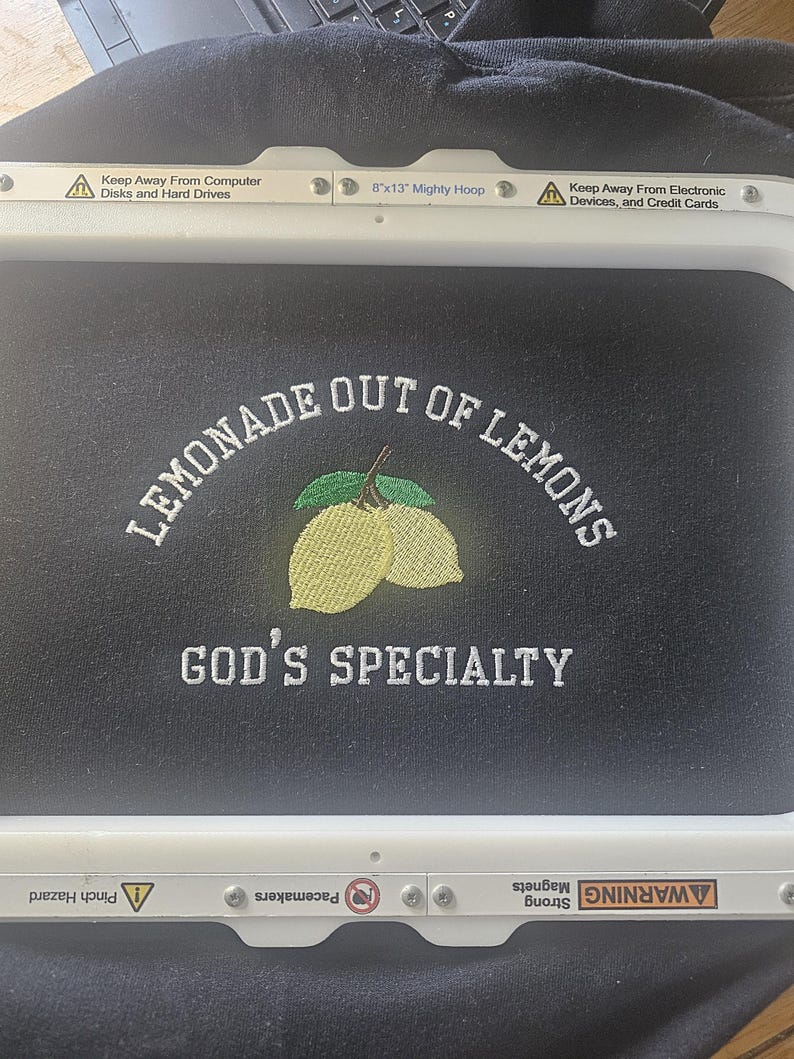 May include: Black fabric with embroidered text and a lemon design. The text reads "LEMONADE OUT OF LEMONS" above "GOD'S SPECIALTY". The design includes two yellow lemons and green leaves. The embroidery is framed by a white hoop.