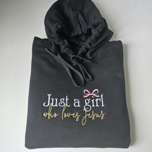 Op de afbeelding: Zwarte hoodie met de geborduurde tekst "Just a girl who loves Jesus". Het ontwerp bevat een roze strik en tekst in wit en goud. De hoodie heeft een trekkoord en een capuchon.
