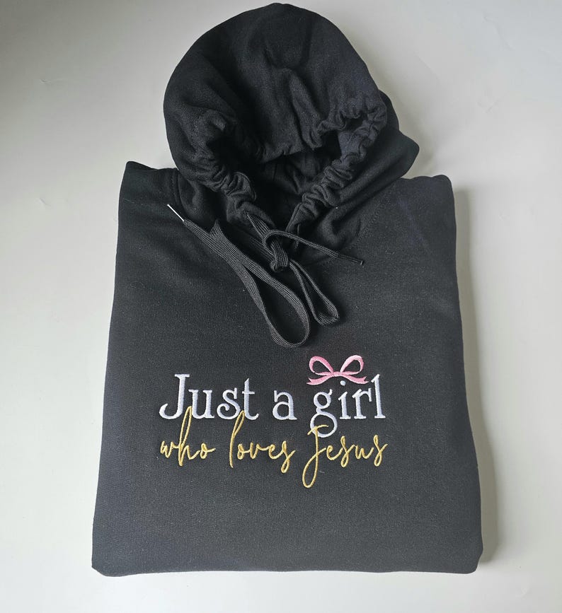 Op de afbeelding: Zwarte hoodie met de geborduurde tekst "Just a girl who loves Jesus". Het ontwerp bevat een roze strik en tekst in wit en goud. De hoodie heeft een trekkoord en een capuchon.
