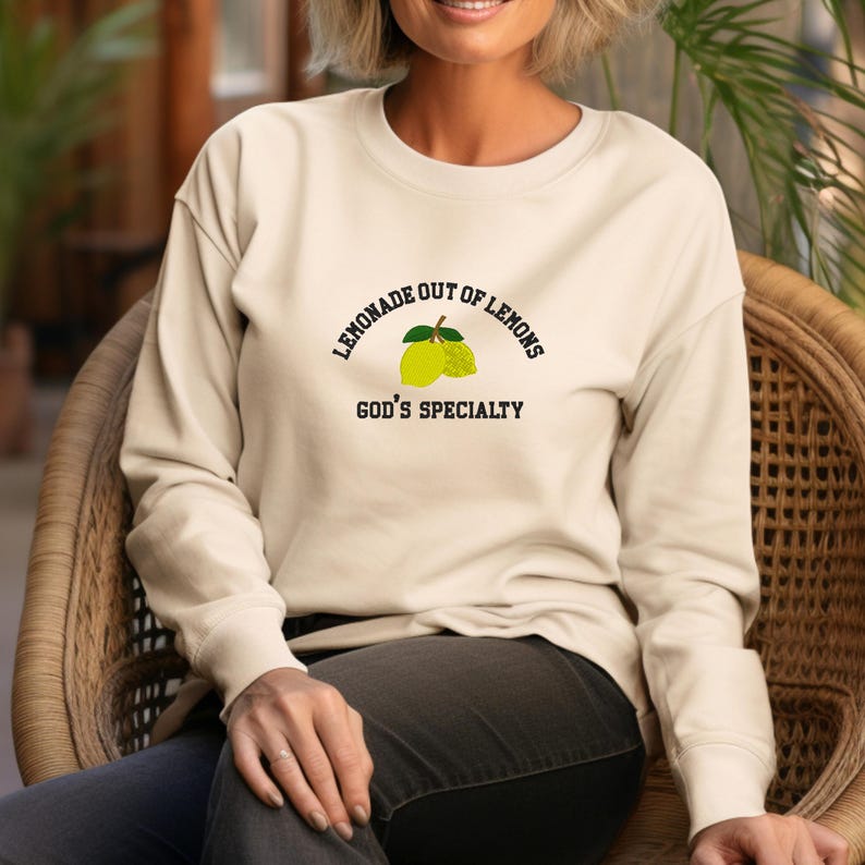 May include: Cream-colored sweatshirt with the text "LEMONADE OUT OF LEMONS" above a lemon graphic and "GOD'S SPECIALTY" below. The long-sleeved top is made of a soft material and is suitable for casual wear.
