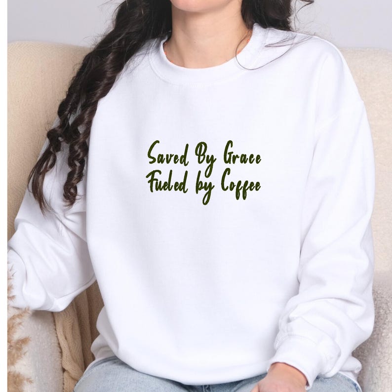 May include: White sweatshirt with the text "Saved By Grace Fueled by Coffee" in a dark green script. The sweatshirt is a solid white color and has a crew neck.