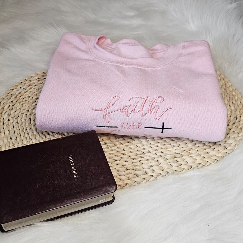 May include: A pink sweatshirt with the text "faith over fear" embroidered on the front. A brown leather-bound Holy Bible is in the foreground.