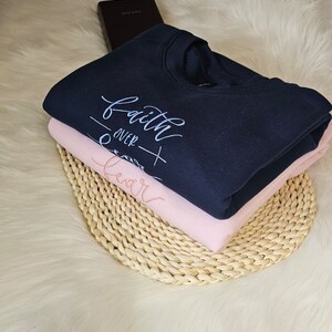 May include: A navy blue sweatshirt with the text "faith over fear" in white embroidery. The sweatshirt is folded and stacked on top of a pink sweatshirt. Both sweatshirts are on a woven straw mat.