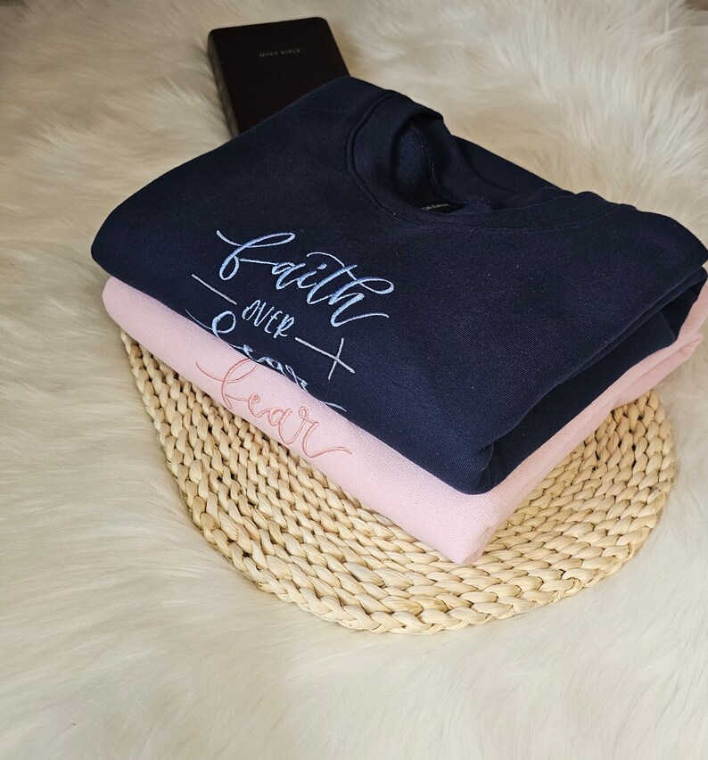 May include: A navy blue sweatshirt with the text "faith over fear" in white embroidery. The sweatshirt is folded and stacked on top of a pink sweatshirt. Both sweatshirts are on a woven straw mat.