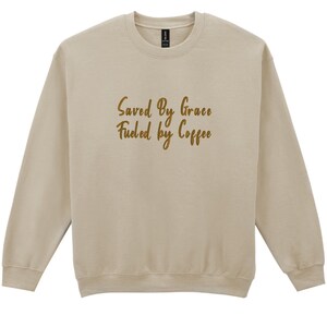 May include: A beige sweatshirt with long sleeves and a crew neck. The text "Saved By Grace Fueled by Coffee" is printed in a gold script font across the front. The sweatshirt is made of a soft material.