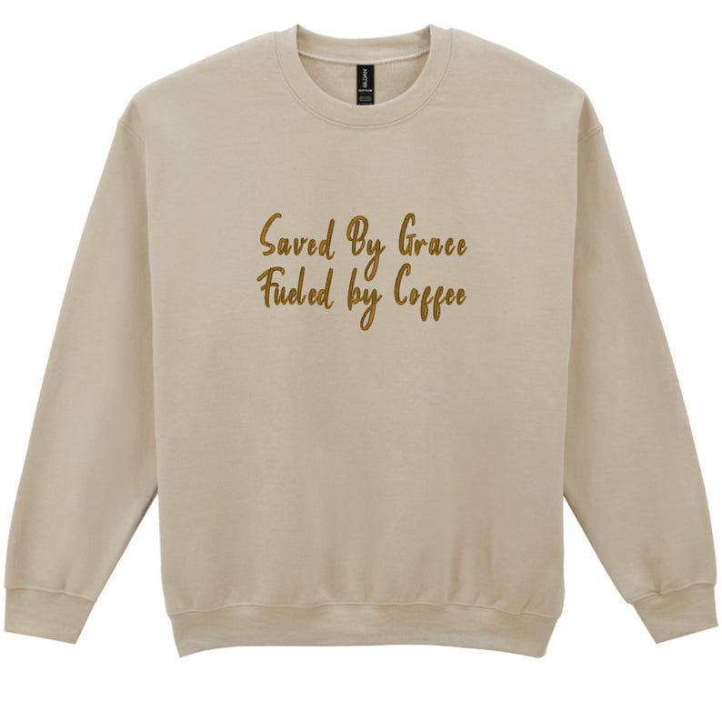 May include: A beige sweatshirt with long sleeves and a crew neck. The text "Saved By Grace Fueled by Coffee" is printed in a gold script font across the front. The sweatshirt is made of a soft material.