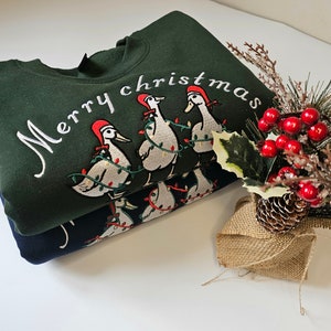 May include: A green sweatshirt with the text "Merry Christmas" embroidered on the front. The sweatshirt features a design of five geese wearing Santa hats and carrying Christmas lights.