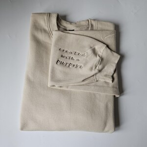 K&ouml;nnte beinhalten: Ein beiger Pullover mit dem gestickten Text "created with a purpose" auf dem &Auml;rmel.