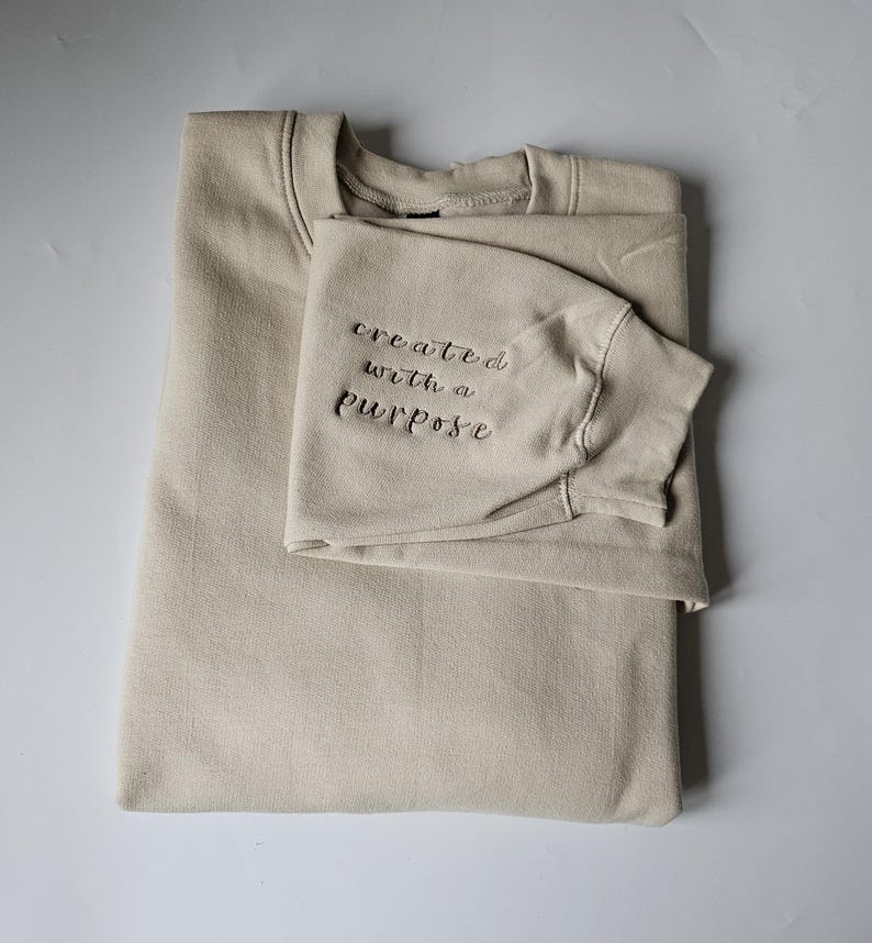 K&ouml;nnte beinhalten: Ein beiger Pullover mit dem gestickten Text "created with a purpose" auf dem &Auml;rmel.
