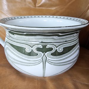 May include: A white ceramic chamber pot with a handle, featuring a decorative design in shades of green, gray, and white. The design includes stylized floral and geometric patterns. The pot is sitting on a brown leather surface.