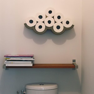 May include: A cloud-shaped toilet paper holder made of gray material, holding several rolls of white toilet paper. Below, a wooden shelf with metal brackets holds a stack of magazines. The scene is set in a bathroom.