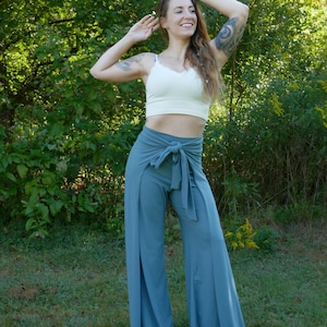 Op de afbeelding: Een vrouw draagt een wit crop top en een blauwe wijde broek met een strik in de taille. De broek heeft aan elke kant een split. Ze staat in een grasachtig gebied met bomen op de achtergrond.