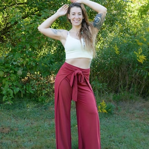 Puede incluir: Una mujer con un top blanco corto y pantalones marrones de estilo envolvente con una cinta ancha. Est&aacute; de pie en una zona cubierta de hierba con &aacute;rboles en el fondo.