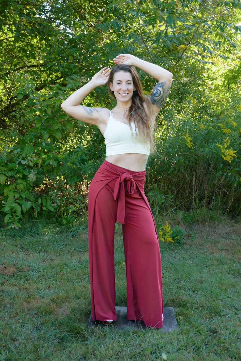 Puede incluir: Una mujer con un top blanco corto y pantalones marrones de estilo envolvente con una cinta ancha. Est&aacute; de pie en una zona cubierta de hierba con &aacute;rboles en el fondo.