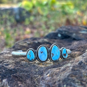 May include: A silver cuff bracelet featuring four oval turquoise stones. Each stone is set in a beaded silver bezel. The turquoise stones have a vibrant blue color with brown matrix patterns. The bracelet is displayed on a textured rock surface.