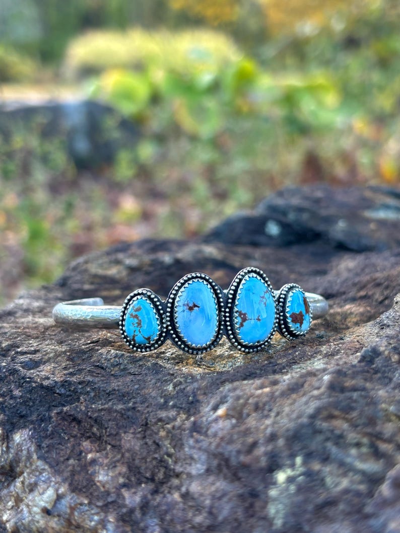 May include: A silver cuff bracelet featuring four oval turquoise stones. Each stone is set in a beaded silver bezel. The turquoise stones have a vibrant blue color with brown matrix patterns. The bracelet is displayed on a textured rock surface.