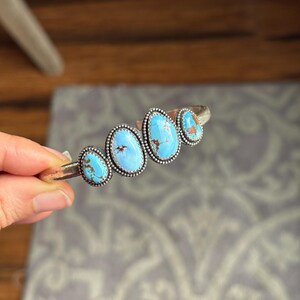 May include: A silver cuff bracelet featuring four oval turquoise stones. The stones are a vibrant blue with brown matrix patterns, set in a beaded silver bezel. The bracelet is held against a neutral background.