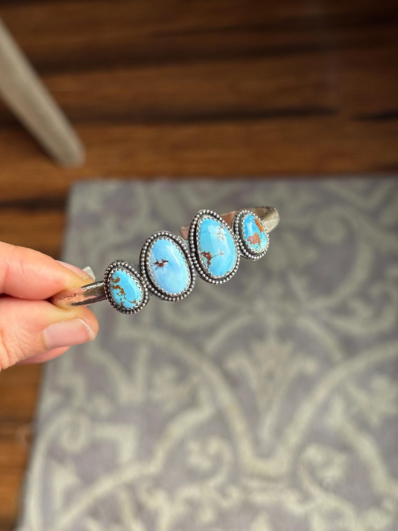 May include: A silver cuff bracelet featuring four oval turquoise stones. The stones are a vibrant blue with brown matrix patterns, set in a beaded silver bezel. The bracelet is held against a neutral background.