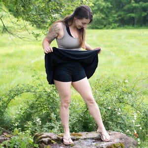 Op de afbeelding: Een zwarte skort met ingebouwde shorts en een neutraal gekleurde tanktop. De skort is uitlopend en de shorts zijn zichtbaar eronder. De persoon staat op een met mos bedekte rots in een groene buitenomgeving.